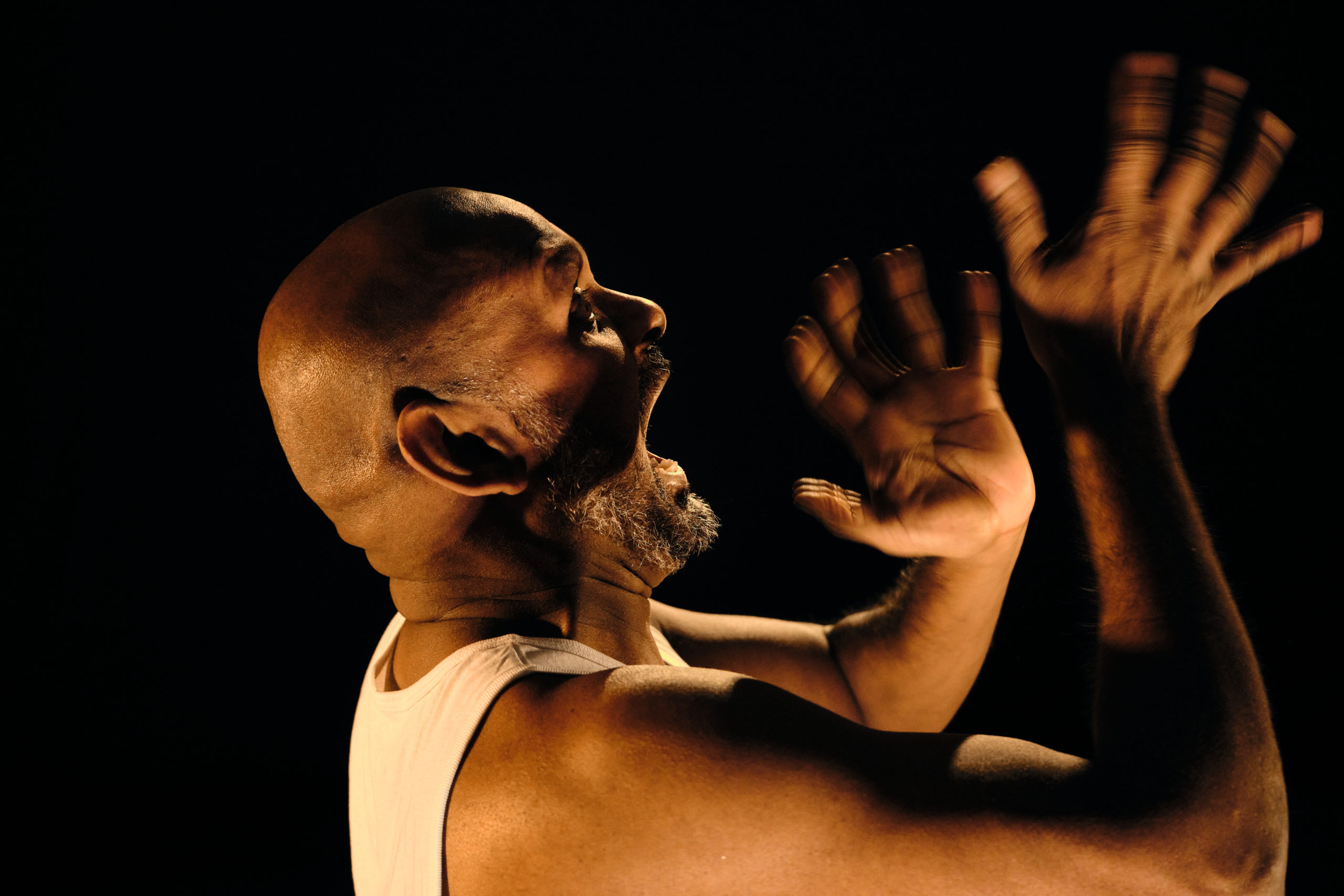 Photo of actor Ramesh Meyyappan performing in Last Rites. A close up of his head and shoulders with his arms raised. His hands are blurred as they are captured in the middle of fast movement back and forwards. His fingers are spread wide apart and his mouth is wide open. Photo by Jack Offord