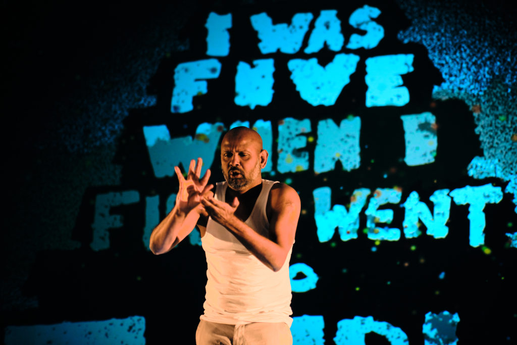 Photo of actor Ramesh Meyyappan performing in Last Rites. He is standing in front of a backdrop which has the words ‘I was five when I first went to temple’ projected in turquoise letters. Ramesh is in the middle of signing and standing in the centre.