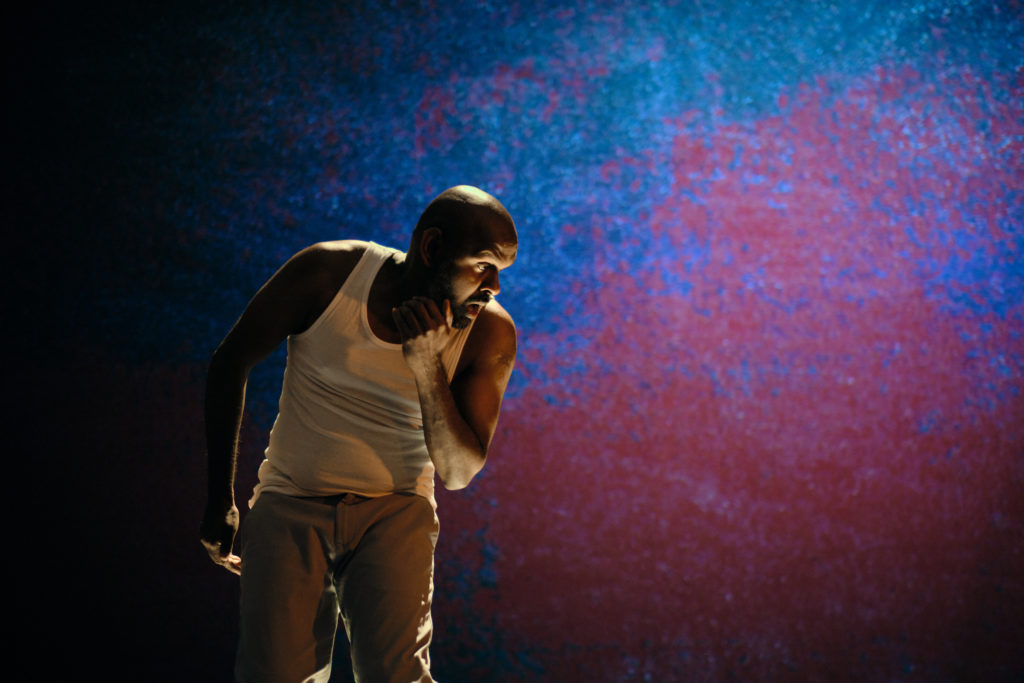 Photo of actor Ramesh Meyyappan performing in Last Rites. He is in front of a backdrop with pink and turquoise projections on it. He is leaning over towards the left and has his hand raised under his chin.